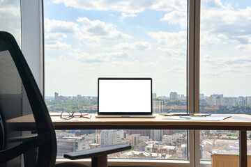 Workplace with laptop on desk with blank empty mockup screen for advertising standing in modern contemporary office on high floor with panoramic big city urban view. No people. Business concept.