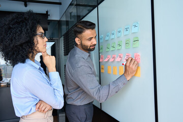 Young indian businessman manager writing strategy ideas on sticky notes on whiteboard and female...