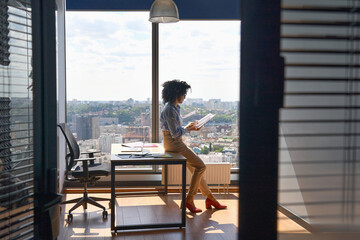Young successful female African American executive manager ceo sitting on desk reading financial report in modern office in high floor corporate building with panoramic urban megapolis city view. - obrazy, fototapety, plakaty
