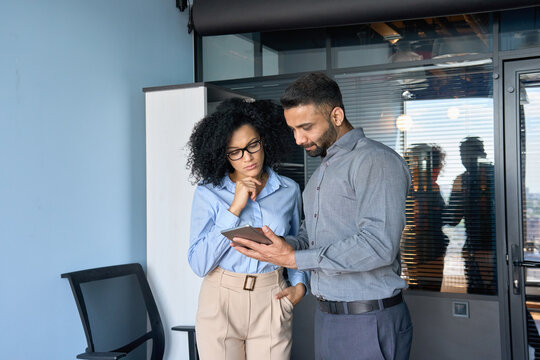 Concentrated Indian male ceo businessman and female African American financial manager working on investment operations using tablet device standing in modern corporate office.