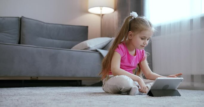 Curious cute preschool kid girl using digital tablet technology device lying on carpet floor alone. Small child hold pad computer surfing internet play game at home. Children tech addiction concept
