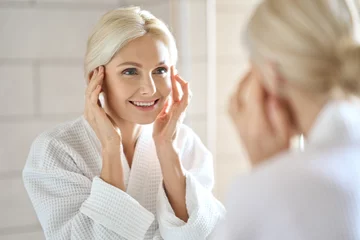 Vlies Fototapete Schönheitssalon Gorgeous mid age adult 50 years old blonde woman standing in bathroom after shower touching face, looking at reflection in mirror smiling doing morning beauty routine. Older dry skin care concept.