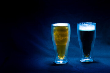 Two glasses of cold and refreshing light and dark beer on a blue cloth and a blue background with a dim light on them