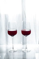 Two crystal glasses with red wine on a glass table and soft backlighting in a living room window