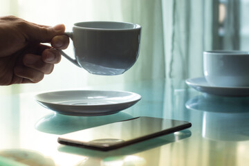a hand sipping a cup of coffee and another cup of coffee on a glass table next to a smartphone with a window with a curtain in the background with dim light
