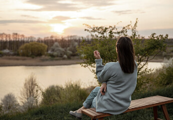 Fototapeta premium A girl in casual style sits and looks at the sunset near the lake.
