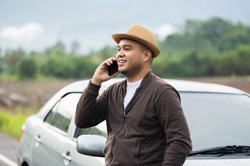 A young Asian man drives a beautiful nature drive. He was standing in front of the car on the roadside. He uses the phone to call services.