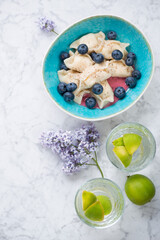 Cottage cheese dumplings with bilberries in a turquoise bowl, flatlay on a light-grey marble background, vertical shot with copy space