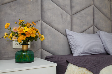 Beautiful grey bedroom, bed, pillows, soft fabric headboard, autumn bouquet of yellow chrysanthemums in green vase near. Modern interior design