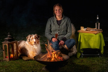 Young woman drinks red wine by the campfire in the forest. Her Australian shepperd sits next to her on a rug in the snowy grass. Enjoying food and drink in leisure time in winter