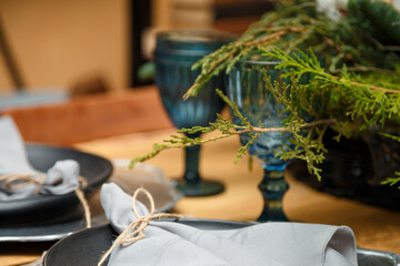 Soft focused shot of rustic style Christmas festive table, blue glasses, plates and fresh thuja branches bouquet. Eco-friendly green decorative elements.