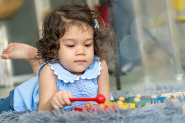 The curly-haired daughter laying on the rug playing with toys, using a bat to play happy tunes in her living room. 