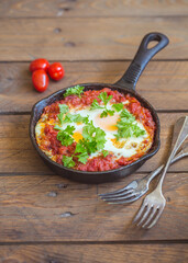Shakshuka - fried eggs with vegetables in iron cast on wooden table