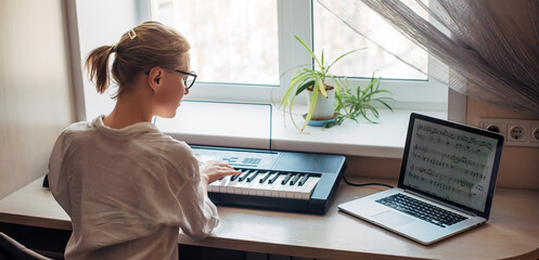 View from the back young woman plays a synthesizer, reading notes on a laptop screen. Independent...