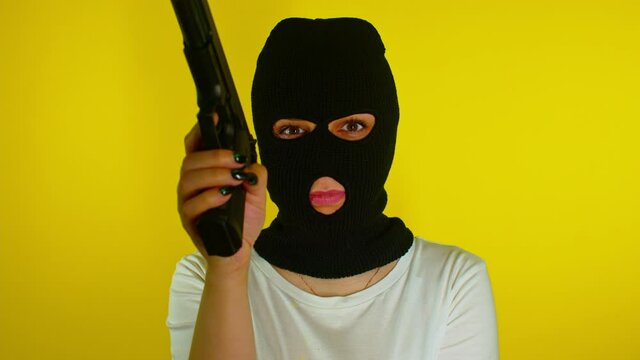 Unrecognizable woman in black balaclava with gun on yellow background. Dangerous female in mask takes aim at camera and shoots. Slow motion