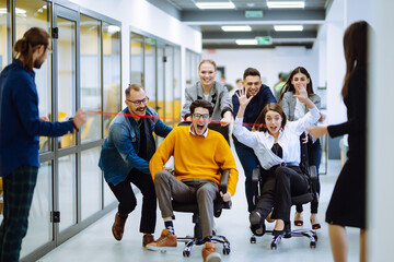 Young colleagues group having fun together, riding on chairs in office, diverse excited office workers enjoying break, laughing, engaged funny activity, celebrating corporate success.