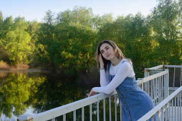 Naklejka na ściany i meble Beautiful young woman stands near railing on the bridge over river. Romantic summer mood.