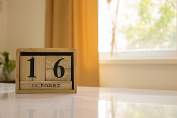 Wooden blocks of the calendar represents the date 16 and the month of October on the background of a window, curtain and a plant.