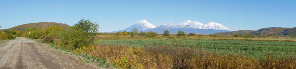 Obraz premium Panorama view of the road and domestic volcanoes against the background of the sky and autumn forest. In good weather, the snow caps of all domestic volcanoes are visible. Kamchatka Peninsula, Russia.