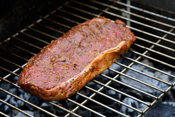 1 raw steak (rump steak) of beef lies on a stainless steel grill grate. Glowing coals roast the delicacy. View from diagonally in front.