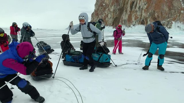 A group of tourists should take shelter from the strong hurricane wind at the foot of the cliff during a winter hike with sledges loaded with backpacks. Storm on Lake Baikal in winter.