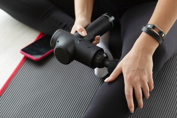 Young fit woman practicing self-massage technique for buttocks applying therapeutic percussive massage gun sitting on pilates mat at home or studio interior.