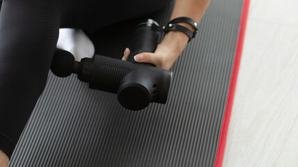 Young fit woman practicing self-massage technique for buttocks applying therapeutic percussive massage gun sitting on pilates mat at home or studio interior.