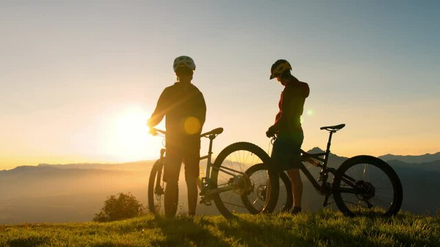 Two female silhouettes,mother and daughter, standing near mountain bikes, talking and observing view in the background, mountaintops