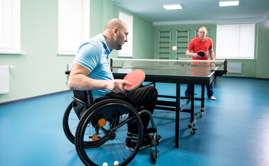 Adult disabled man in a wheelchair play at table tennis with his coach