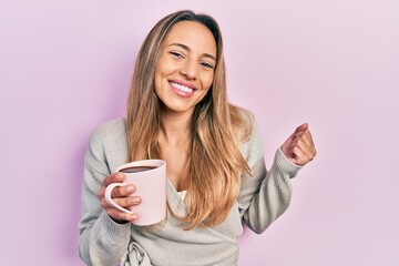 Beautiful hispanic woman drinking a cup coffee screaming proud, celebrating victory and success very excited with raised arm