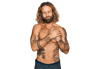 Handsome man with beard and long hair standing shirtless showing tattoos smiling with hands on...