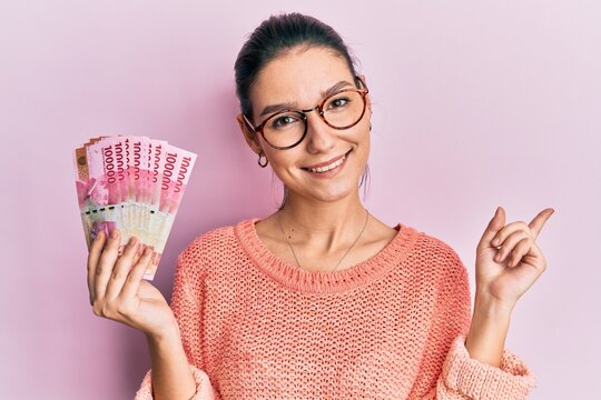 Young caucasian woman holding 100000 indonesian rupiah smiling happy pointing with hand and finger to the side