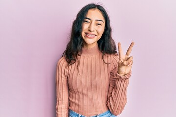 Hispanic teenager girl with dental braces wearing casual clothes smiling with happy face winking at the camera doing victory sign. number two.