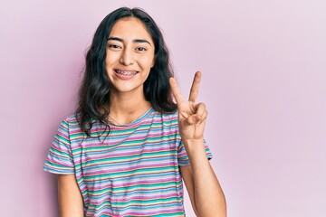 Hispanic teenager girl with dental braces wearing casual clothes showing and pointing up with...