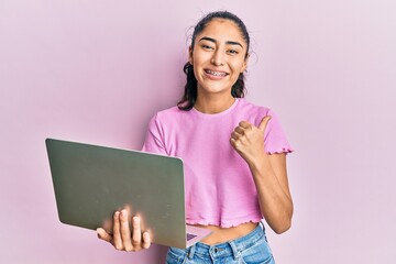 Hispanic teenager girl with dental braces holding and using computer laptop pointing thumb up to...