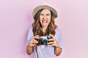 Young hispanic girl wearing summer hat holding vintage camera sticking tongue out happy with funny...