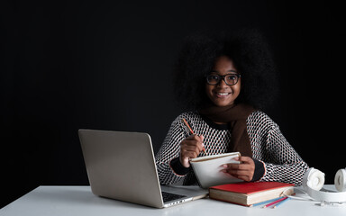 Portrait of African girl wear glasses smiling and writing on book while online learning with laptop textbooks headphones. Teenage girl looking at camera with sweater on black background. Copy space