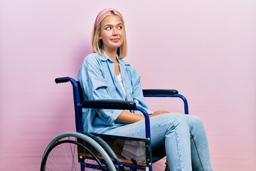 Beautiful blonde woman sitting on wheelchair smiling looking to the side and staring away thinking.