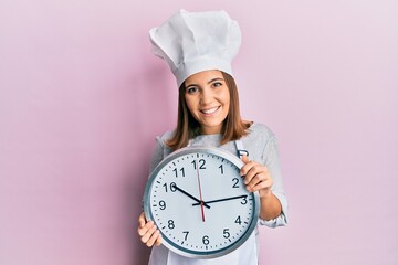 Young beautiful woman wearing professional cook uniform and hat holding clock smiling with a happy...