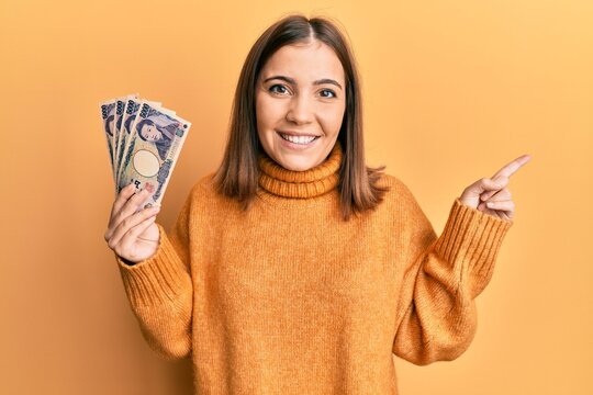 Young woman holding 5000 japanese yen banknotes smiling happy pointing with hand and finger to the side