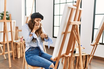 Young hispanic artist woman painting on canvas at art studio looking at the watch time worried,...