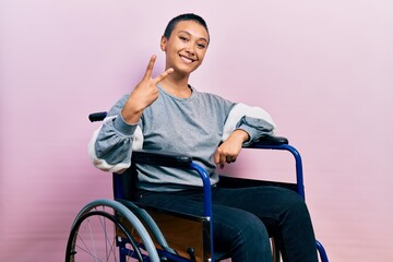 Beautiful hispanic woman with short hair sitting on wheelchair showing and pointing up with fingers...