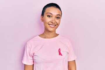 Fototapeta premium Beautiful hispanic woman with short hair wearing pink cancer ribbon on shirt looking positive and happy standing and smiling with a confident smile showing teeth