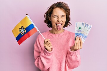 Young brunette woman holding colombia flag and colombian pesos banknotes sticking tongue out happy...