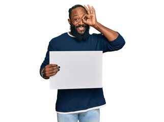 Young african american man holding blank empty banner smiling happy doing ok sign with hand on eye looking through fingers