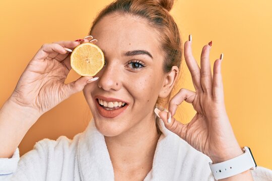 Young blonde woman wearing bathrobe holding lemon over eye doing ok sign with fingers, smiling friendly gesturing excellent symbol