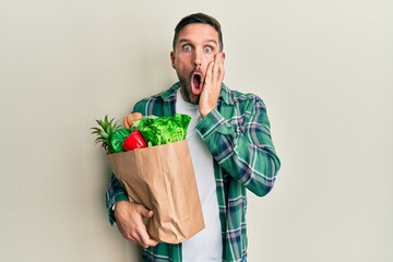 Handsome man with beard holding paper bag with groceries afraid and shocked, surprise and amazed expression with hands on face