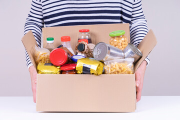 Volunteer female hands holding food donations box with grocery products on white desk. Front view