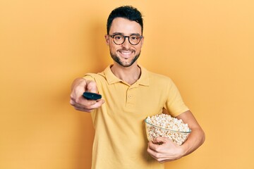 Young hispanic man eating popcorn using tv control smiling with a happy and cool smile on face....