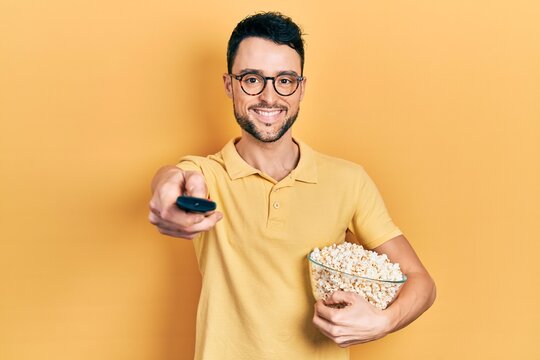 Young hispanic man eating popcorn using tv control smiling with a happy and cool smile on face. showing teeth.